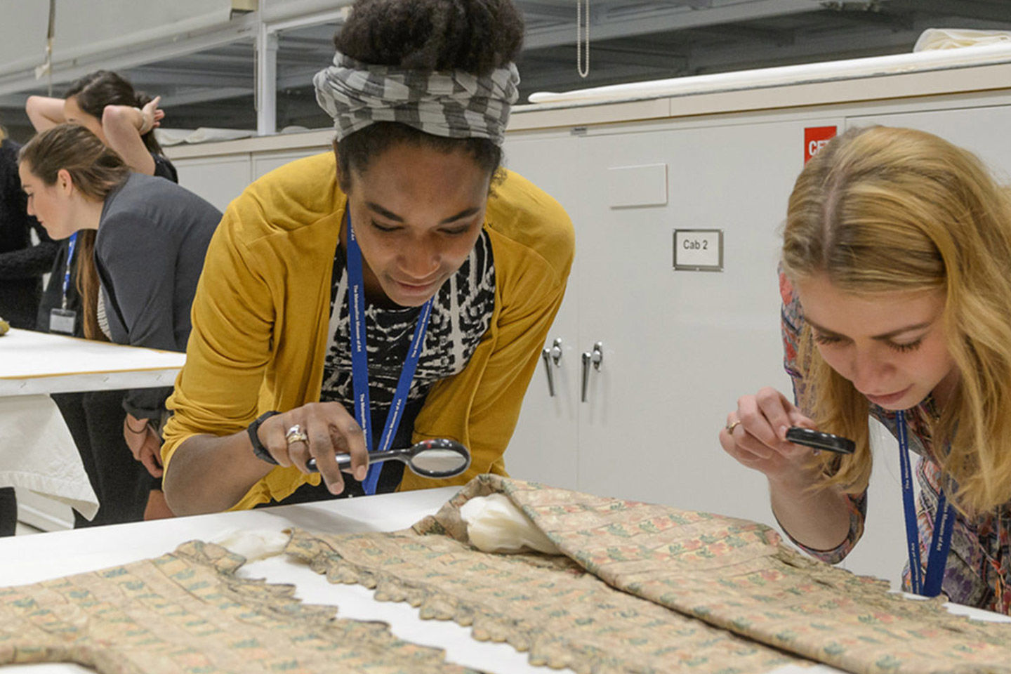Two young women use magnifying glasses to inspect a textile